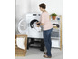 Une femme charge une machine à laver avec un panier à linge à côté.