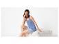 Femme en short et débardeur bleu assise sur un pouf blanc, souriante.