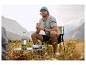 Homme en casquette et t-shirt, assis sur une chaise de camping avec lanterne et table.