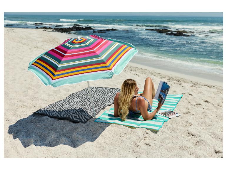 Femme lisant sous un parasol de plage coloré sur un tapis rayé au bord de la mer.