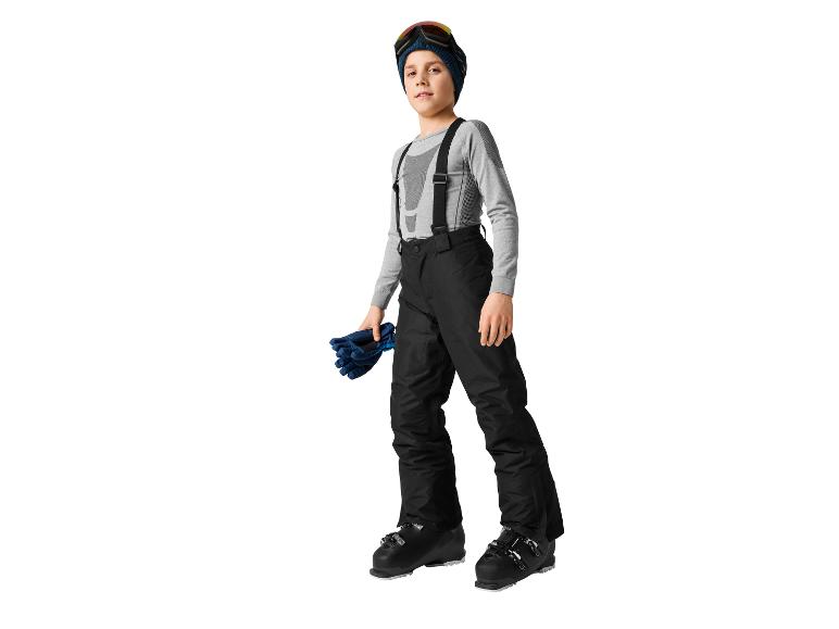 Un jeune garçon en tenue de ski, comprenant un pantalon de ski, un maillot thermique, un bonnet et des lunettes de ski.