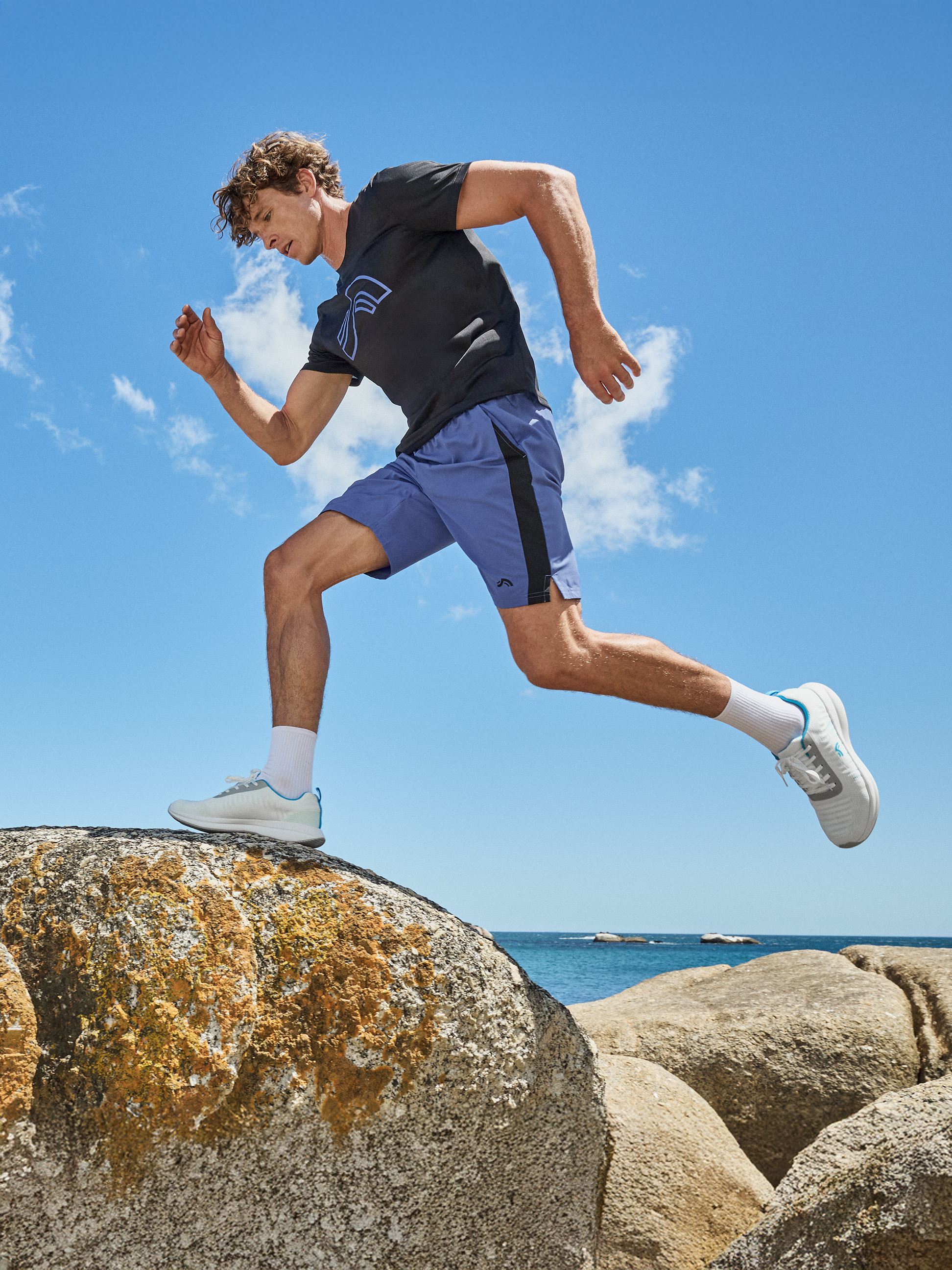 Homme en t-shirt noir et short bleu courant sur des rochers au bord de la plage.
