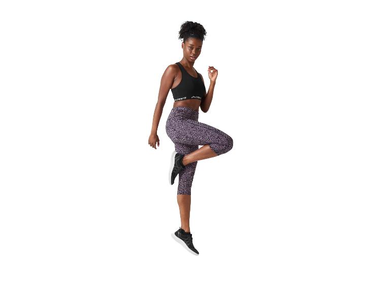 Femme en brassière de sport noire et legging capri imprimé léopard violet, en plein saut.