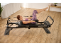 Une femme s'entraîne sur un appareil de pilates reformer noir dans une pièce lumineuse au sol en bois.