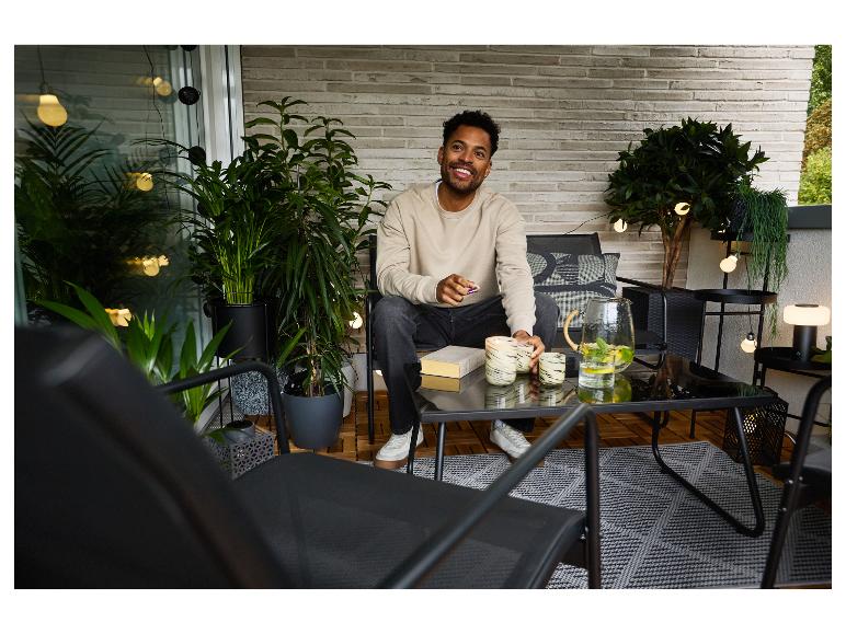 Homme sur balcon avec mobilier d'extérieur, plantes et guirlandes lumineuses.