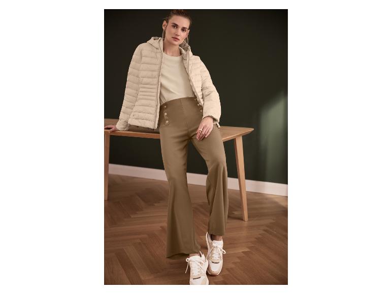 Femme en doudoune beige, t-shirt beige et pantalon marron à boutons, appuyée sur une table.