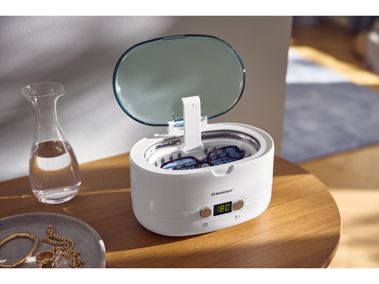 Nettoyeur à ultrasons avec lunettes sur une table en bois, à côté d'une carafe et de bijoux.