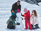 Une famille construit un bonhomme de neige dans la neige, les enfants portent des vêtements d'hiver rouges et verts.
