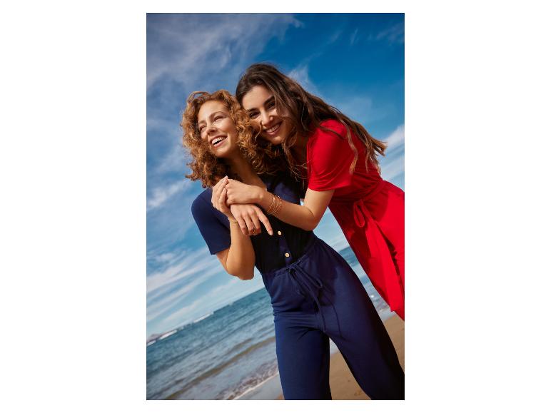 Deux femmes souriantes en combinaisons sur la plage, l'une en bleu marine, l'autre en rouge.