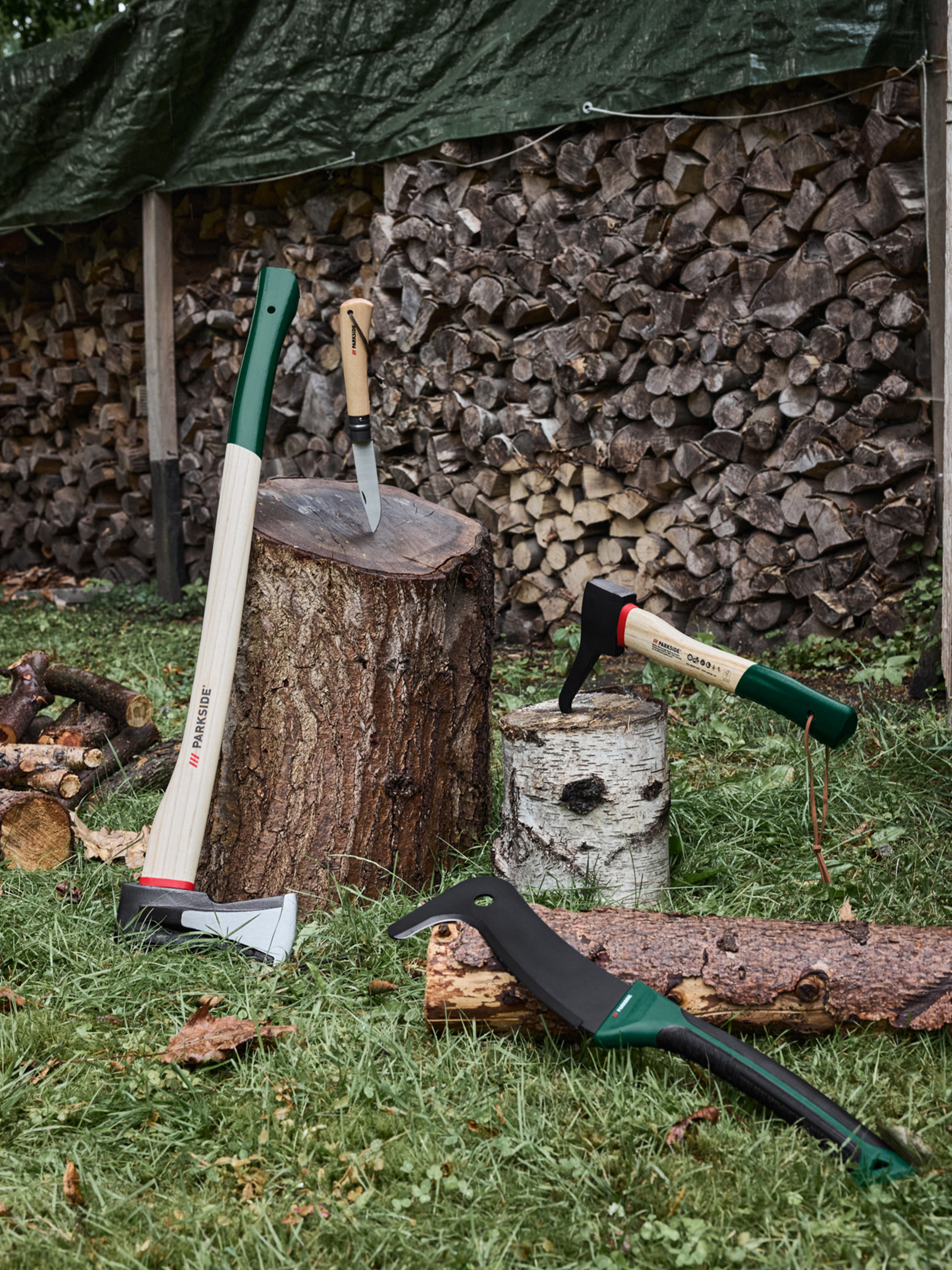 Haches, un couteau et une machette sur l'herbe avec du bois de chauffage empilé en arrière-plan.