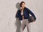 Une femme vêtue d'un sweat à capuche bleu, de leggings gris et tenant un ballon de fitness noir.