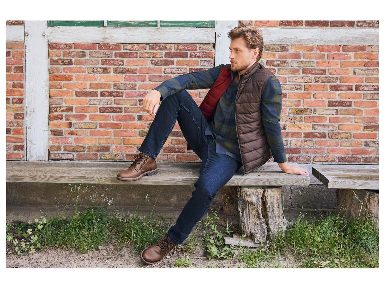 Homme en gilet marron, chemise à carreaux et jean assis sur un banc en bois.