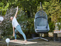Une femme fait du yoga sur un tapis dans le jardin, à côté d'un fauteuil suspendu.