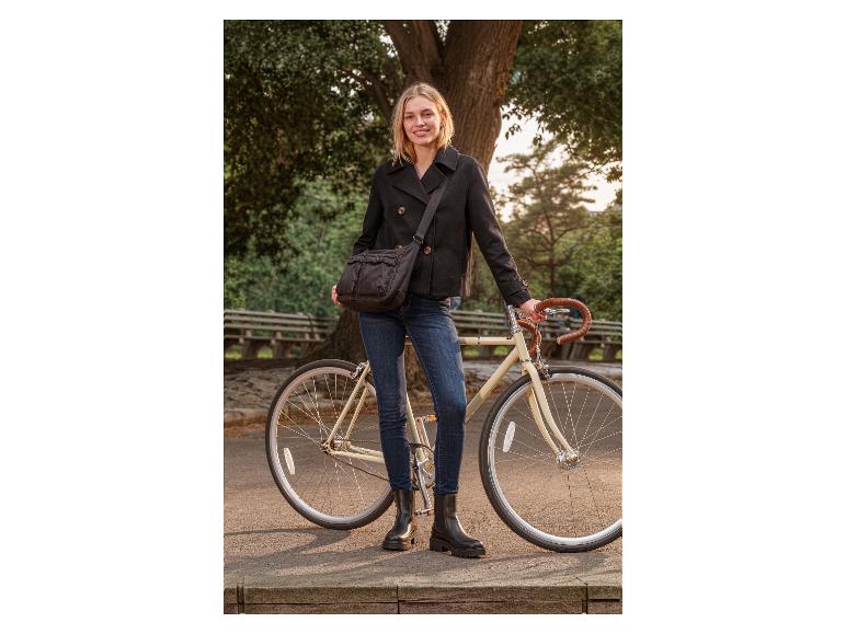 Femme en caban noir et jean avec un sac à bandoulière noir, debout à côté d'un vélo.