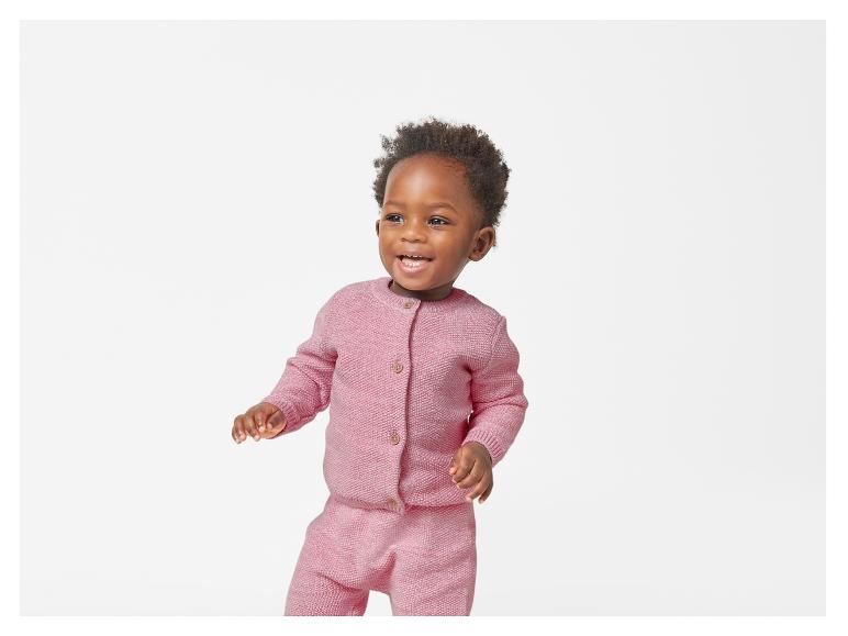 Un jeune enfant sourit en portant un cardigan et un pantalon roses en tricot.