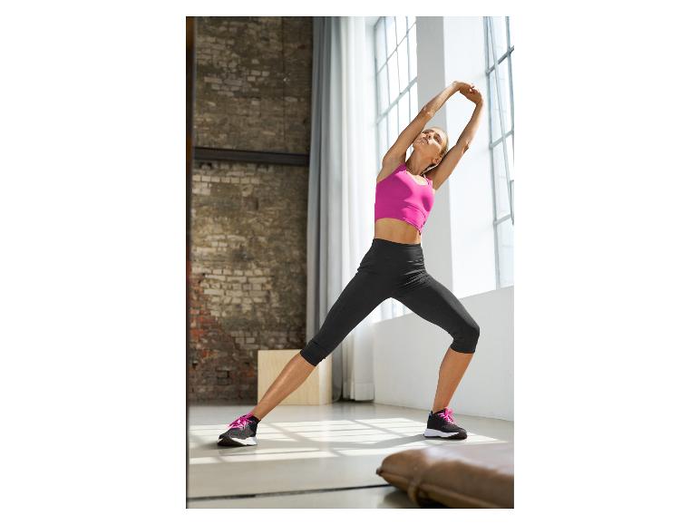 Femme en débardeur de sport rose et legging noir s'étirant dans une pièce lumineuse.