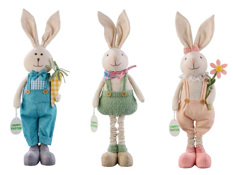 Trois lapins de Pâques en peluche de différentes couleurs avec une carotte, une fleur et une étiquette 'Happy Easter'.