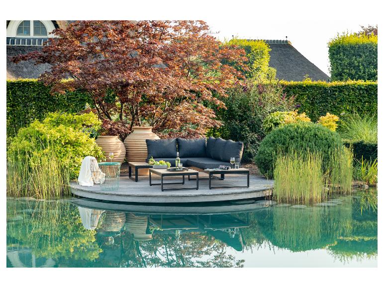 Mobilier de jardin sur une terrasse en bois au bord de l'eau, entouré de verdure luxuriante.