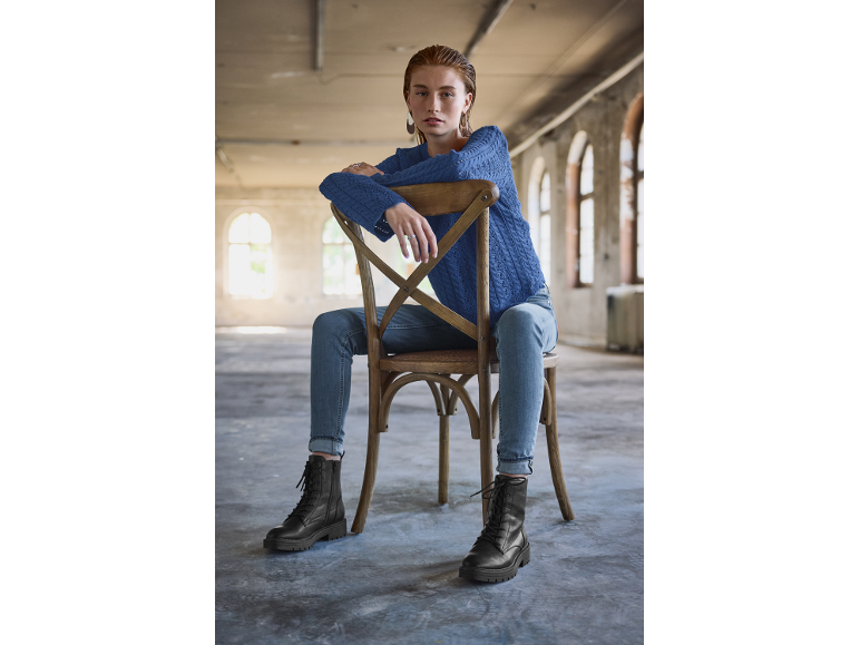 Femme en pull bleu et bottes noires assise sur une chaise en bois.
