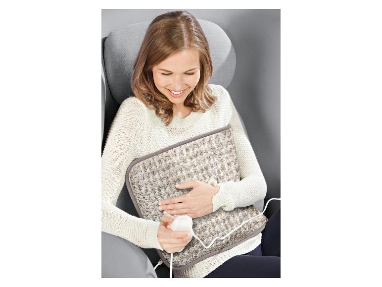 Une femme assise dans un fauteuil sourit en tenant un coussin chauffant électrique beige sur ses genoux.