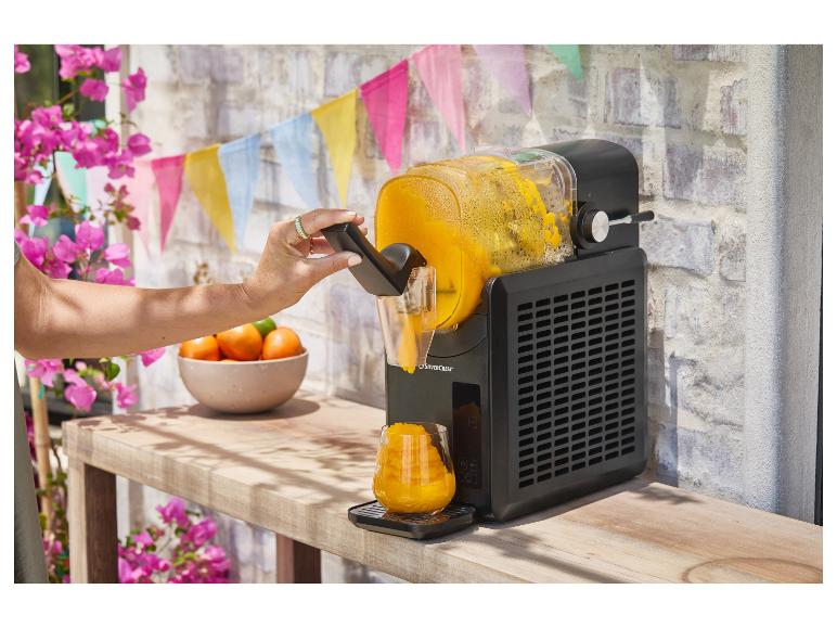 Machine à granita noire servant une boisson à l'orange dans un verre, avec décorations de fête.