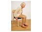 Femme âgée assise sur une chaise en bois avec un coussin noir, vêtue de vêtements beiges.