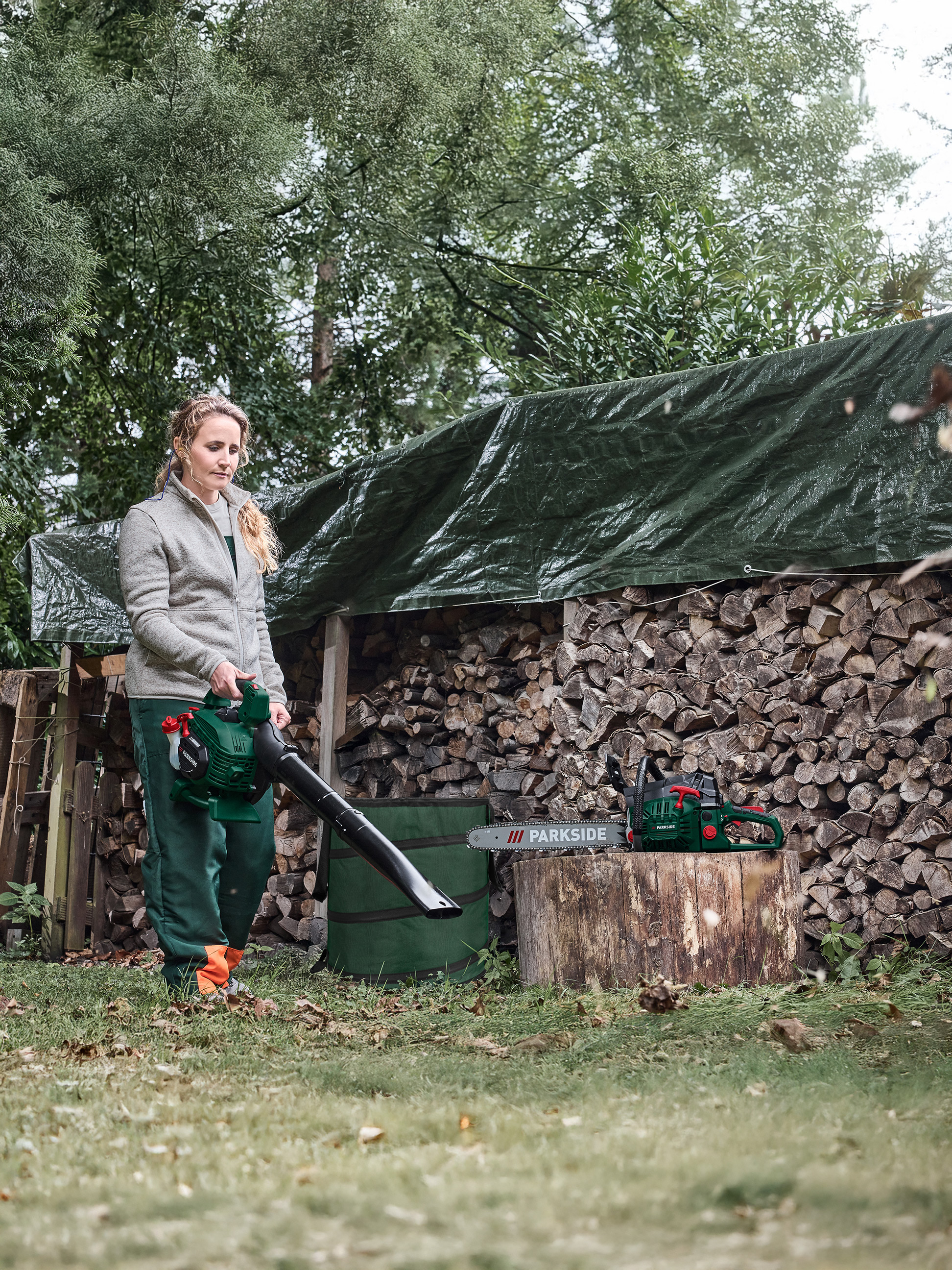 Une femme utilise un souffleur de feuilles près d'un tas de bois de chauffage, avec une tronçonneuse sur une souche.
