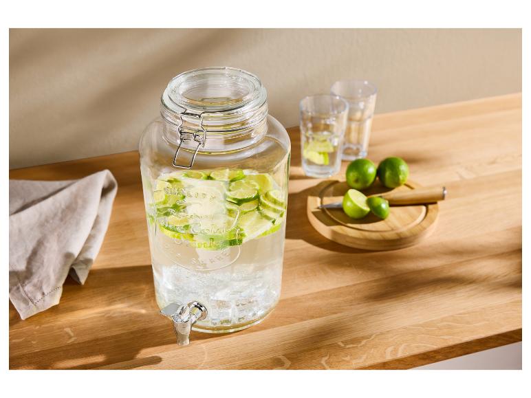 Distributeur de boissons en verre avec des citrons verts tranchés, des verres et des citrons verts entiers sur une table en bois.