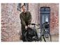 Un homme avec une veste verte regardant un vélo électrique gris avec des sacoches et un casque près d'un mur de briques