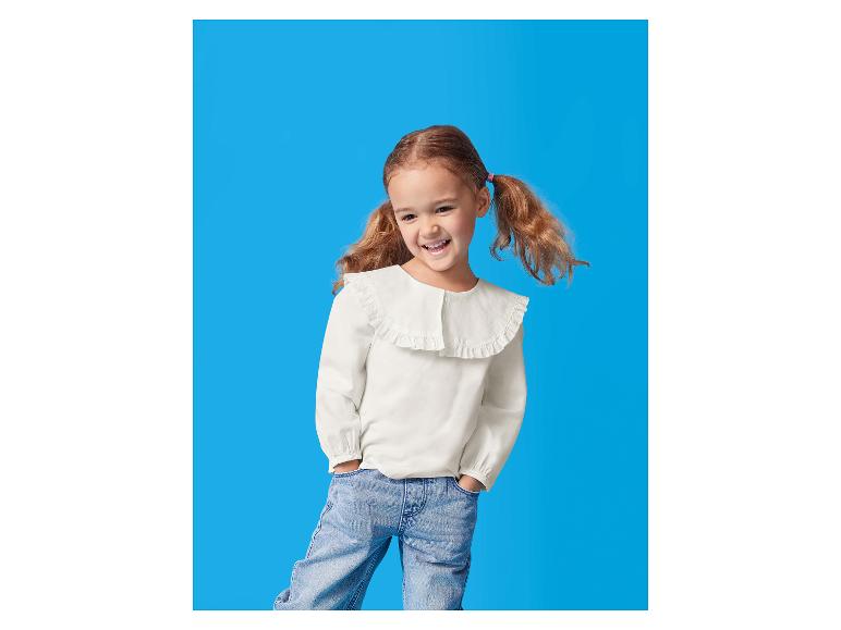 Une fille souriante avec des couettes, portant un chemisier blanc à volants et un jean.