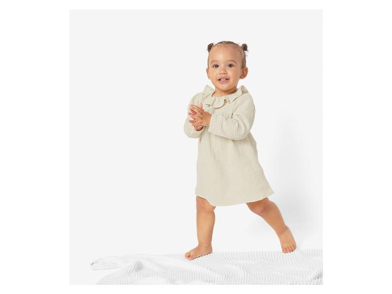 Bébé portant une robe beige et debout sur un tapis blanc.
