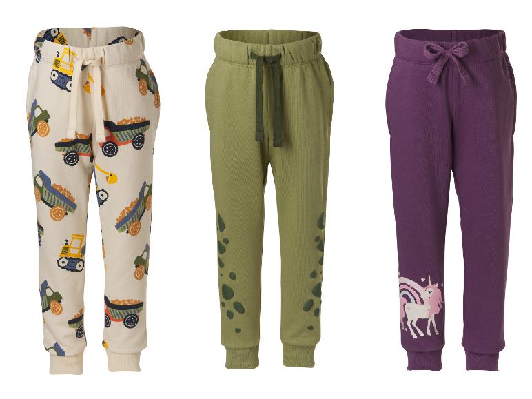 Trois pantalons de jogging pour enfants : un beige à motifs de camions, un vert kaki avec des taches et un violet avec une licorne.