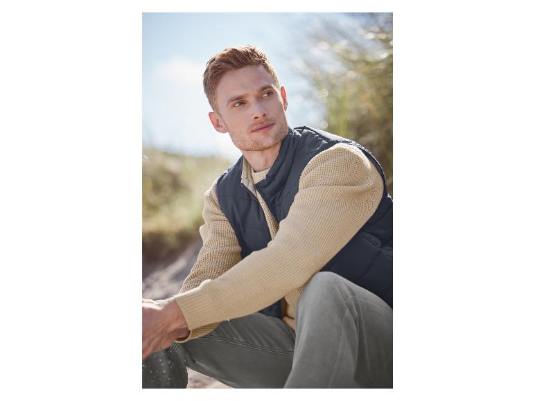 Un homme portant un gilet bleu foncé, un pull beige et un pantalon gris.