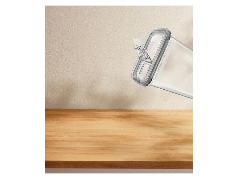 Récipient de conservation alimentaire transparent avec couvercle gris, incliné contre un mur beige et une surface en bois.