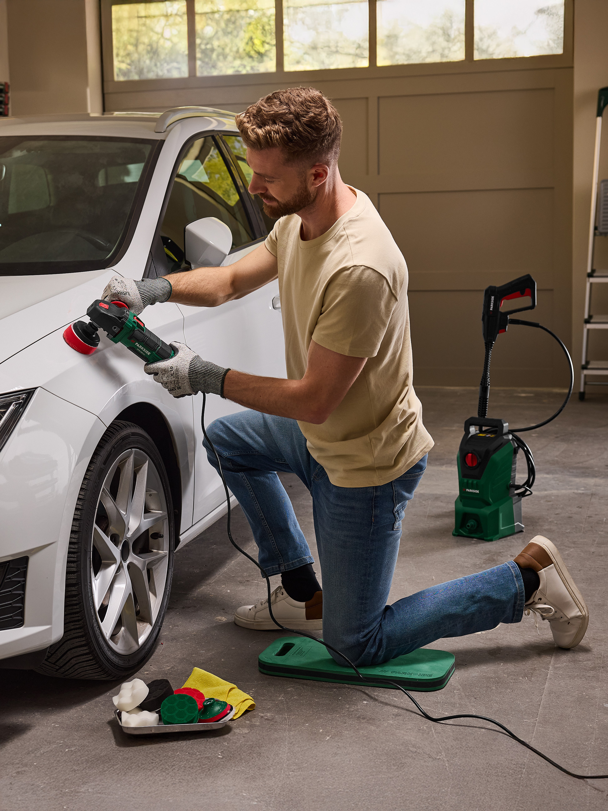 Homme polissant une voiture blanche avec une polisseuse, agenouillé sur un tapis de protection.
