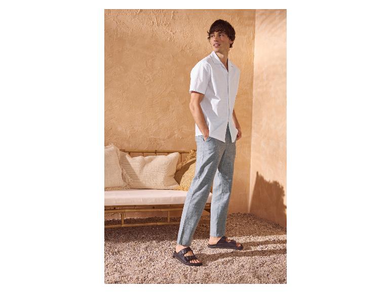 Homme en chemise blanche à manches courtes, pantalon gris et sandales noires dans un décor méditerranéen.