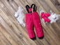 Pantalon de ski rose avec bretelles, mitaines et boules de neige sur un fond en bois.