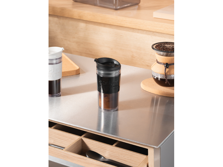Deux tasses de voyage et une cafetière sur un comptoir métallique.