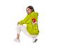 Femme accroupie, portant un sweat à capuche vert Grinch et un pantalon blanc.