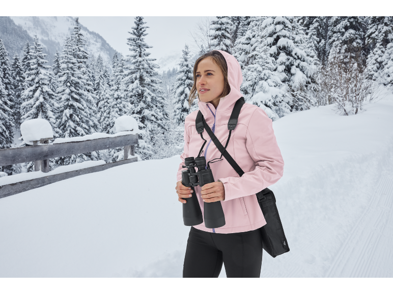 Femme avec des jumelles et une veste rose dans un paysage hivernal enneigé.