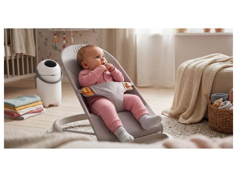 Bébé dans un transat gris, entouré de linge de maison et d'une poubelle à couches.