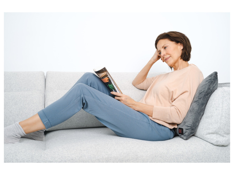 Une femme lit un magazine sur un canapé gris avec un coussin chauffant.