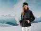 Femme en veste noire et pantalon blanc tenant des skis dans un paysage enneigé.
