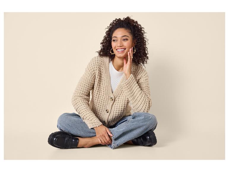 Femme en cardigan tricoté beige, jean et mocassins noirs, assise en tailleur.