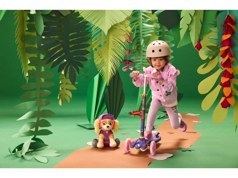 Fille avec un casque sur une trottinette, à côté d'un chien en peluche, dans un décor de jungle.