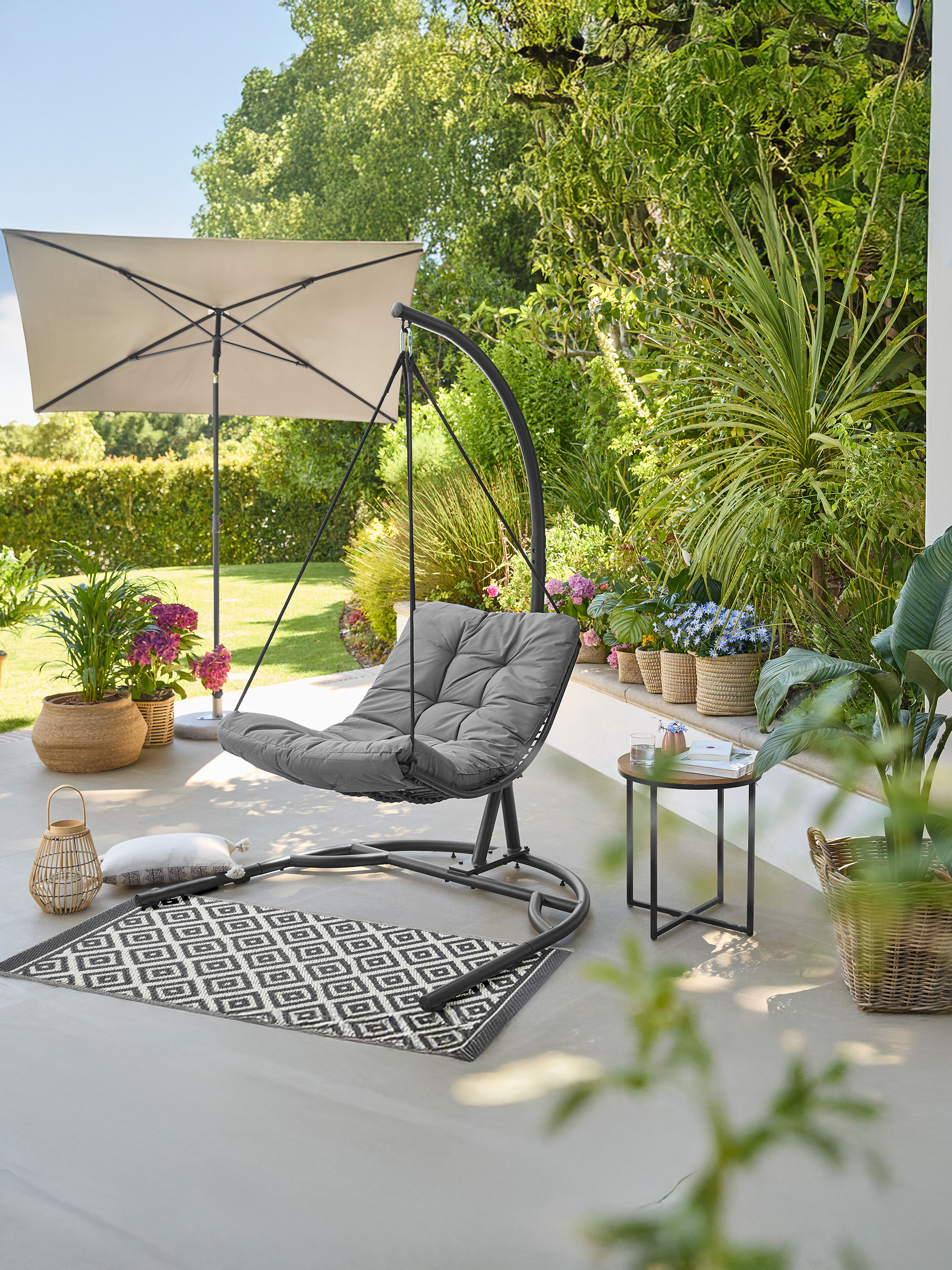 Fauteuil suspendu d'extérieur avec coussin gris, parasol et plantes sur une terrasse.