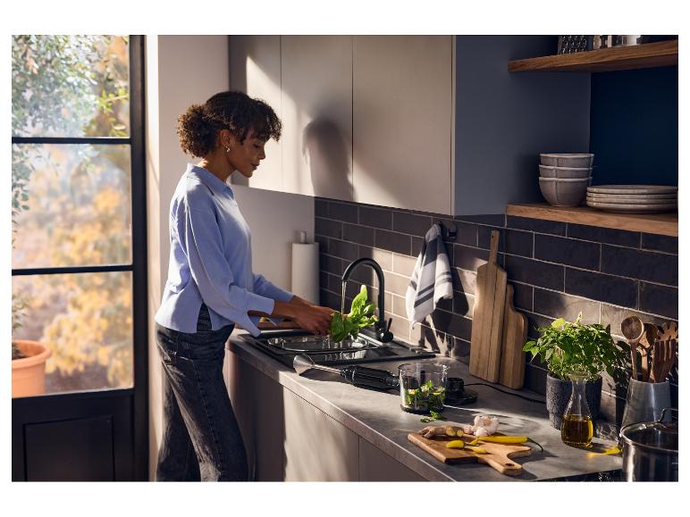 Femme lavant des épinards dans une cuisine moderne avec un mixeur plongeant et des ingrédients sur le comptoir.