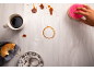 Une main nettoie une table avec une éponge, du café renversé, un bagel et du sucre.