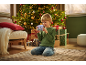 Un garçon agenouillé sur un tapis, tenant un appareil photo jouet, avec un sapin de Noël et des cadeaux derrière lui.