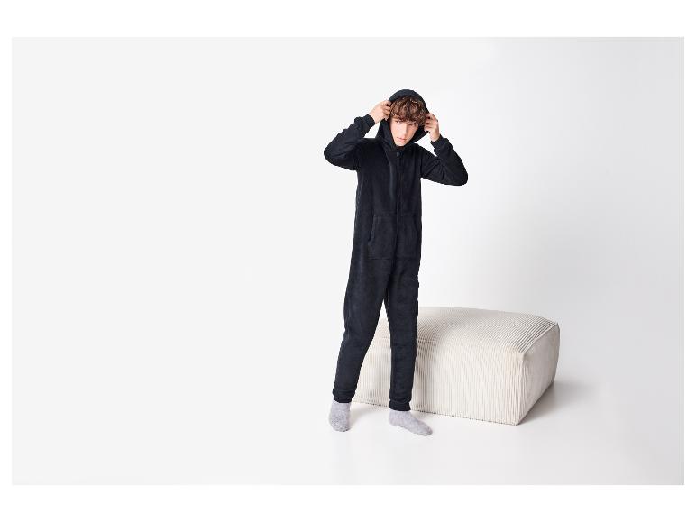 Garçon en combinaison noire à capuche et chaussettes grises, debout près d'un pouf blanc.